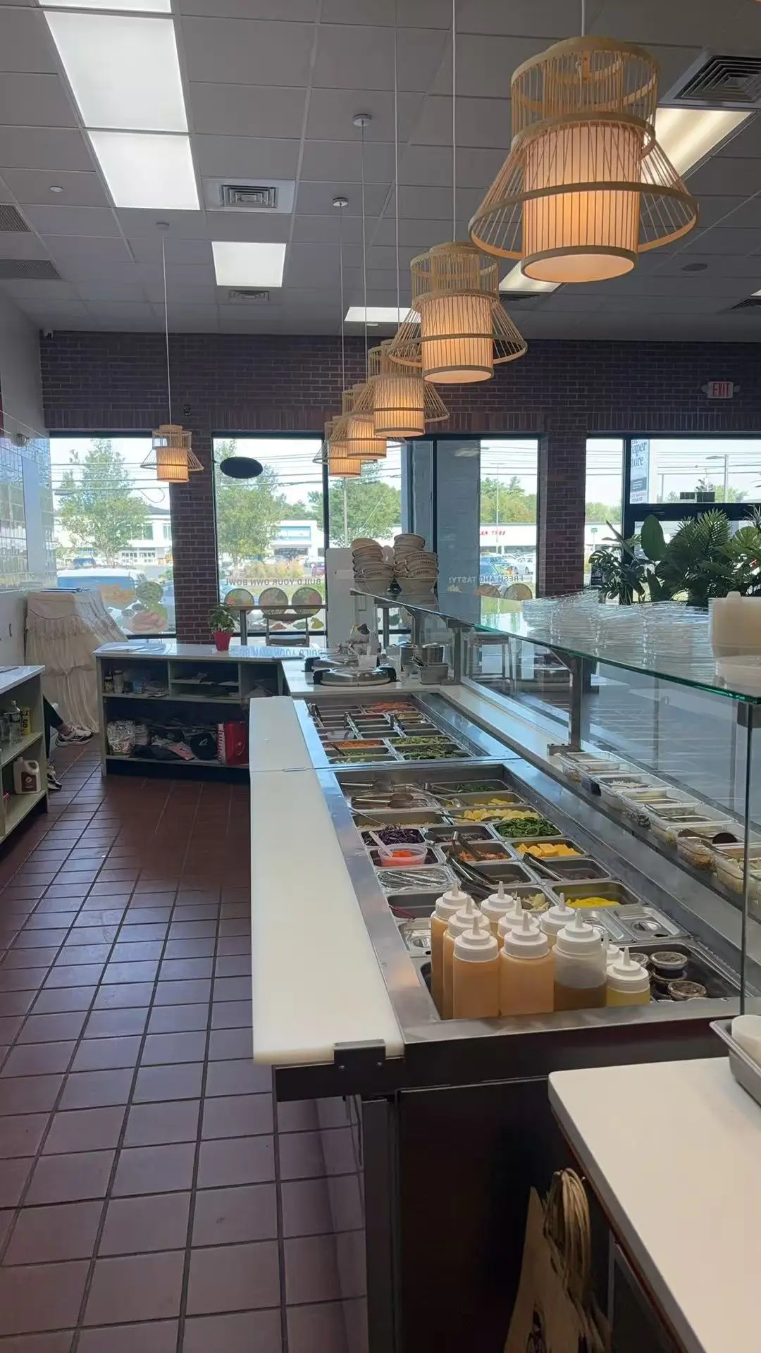 Self-serve poke bar with stainless steel trays of fresh ingredients, sauce bottles, and warm pendant lighting over a tiled floor.